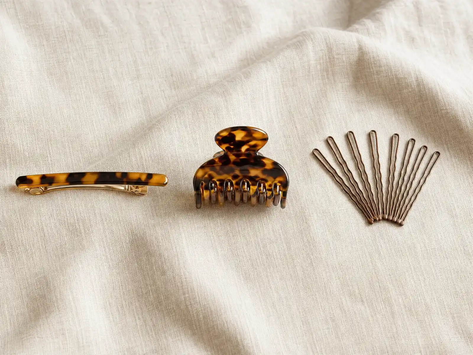 Three hair accessories laid side by side on a linen surface: a slim tortoiseshell barrette with its clasp open, a small nylon claw clip resting on its teeth, and a scattered handful of brown wavy bobby pins