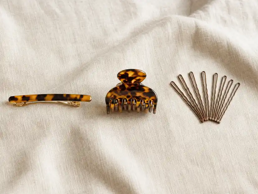 Three hair accessories laid side by side on a linen surface: a slim tortoiseshell barrette with its clasp open, a small nylon claw clip resting on its teeth, and a scattered handful of brown wavy bobby pins