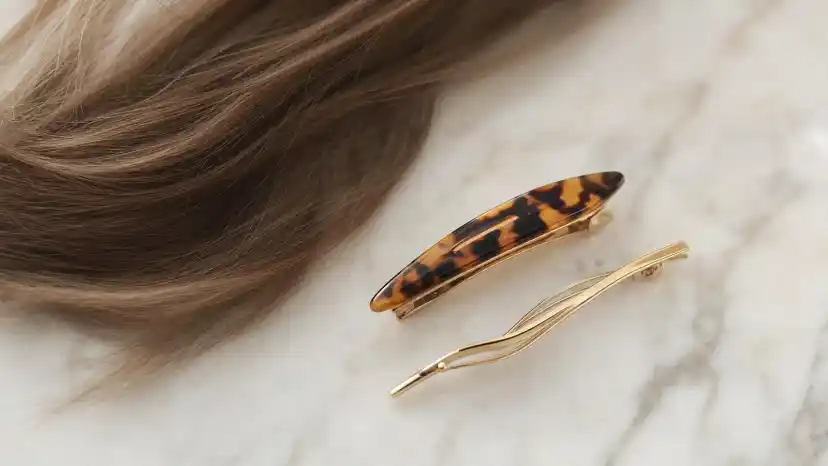 Small tortoiseshell and thin gold barrettes beside a lock of fine light-brown hair on a marble surface