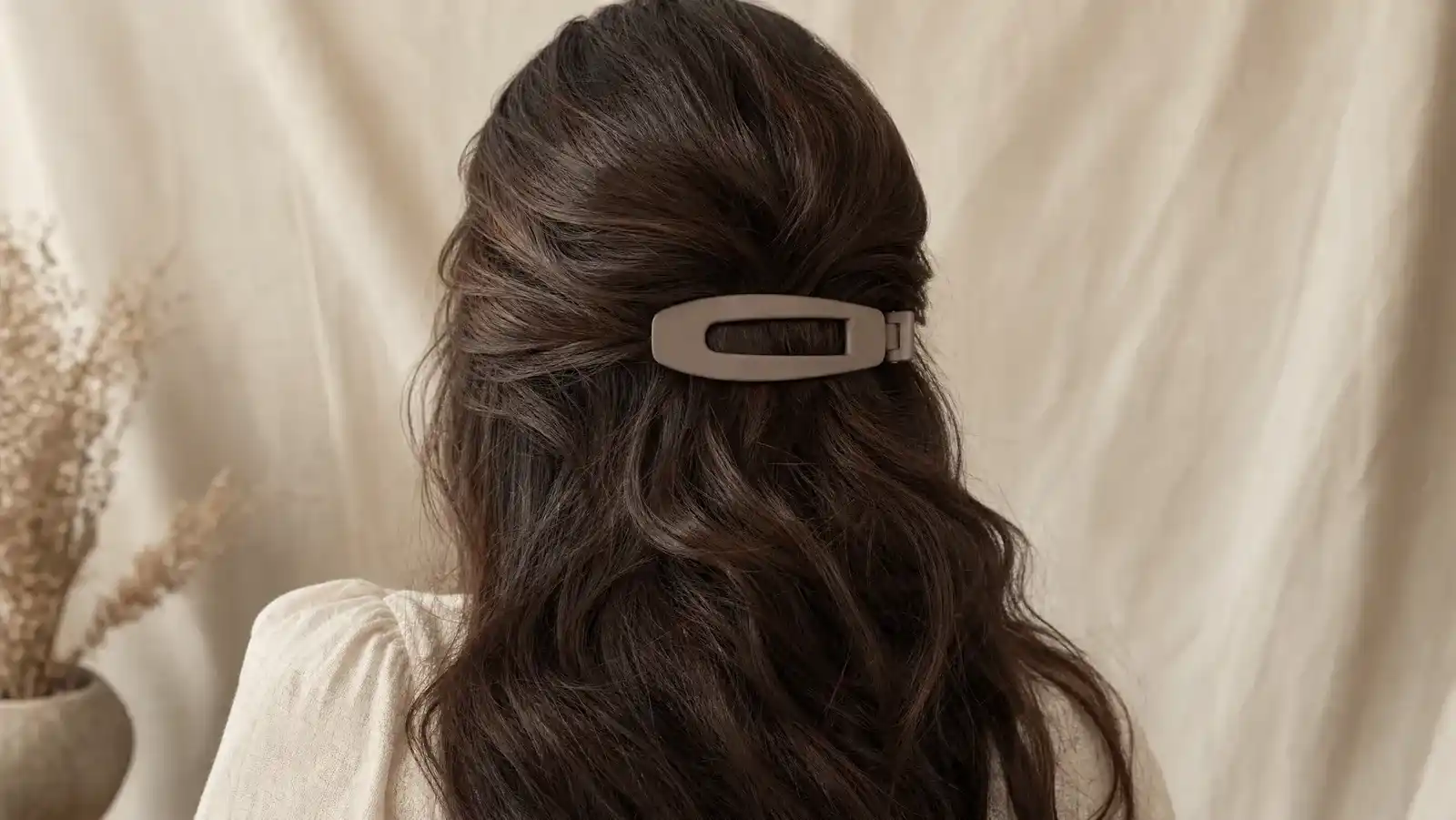 Large matte 4-inch French barrette clasped around a thick section of dark wavy hair, with oversized metal snap clips arranged beside it on a wooden vanity