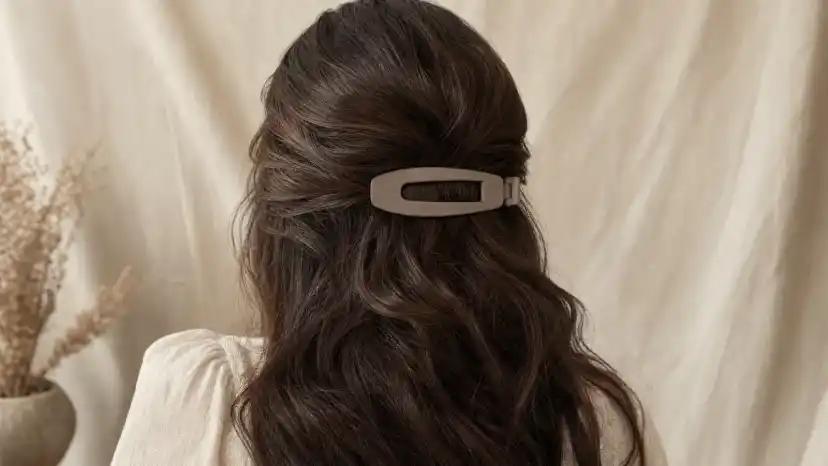Large matte 4-inch French barrette clasped around a thick section of dark wavy hair, with oversized metal snap clips arranged beside it on a wooden vanity