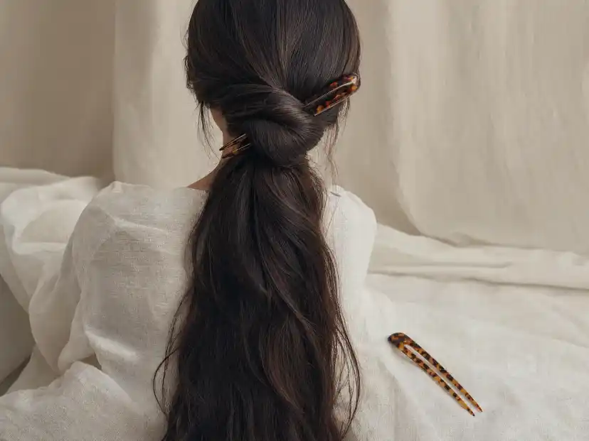 A long tortoiseshell acetate french hair pin crossing a low twisted chignon in waist-length dark brown hair, photographed from behind against a pale linen backdrop with a second pin resting nearby