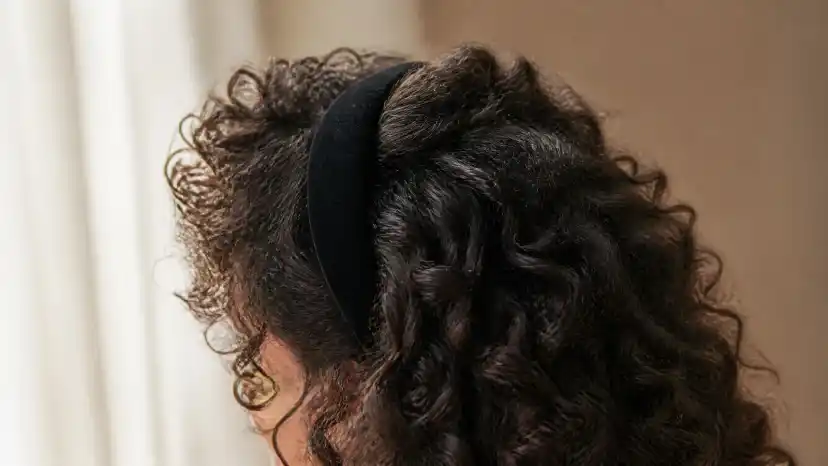 A wide black velvet padded headband set back from the hairline in dark curly hair, with the curl pattern still springing up in front of and behind the band
