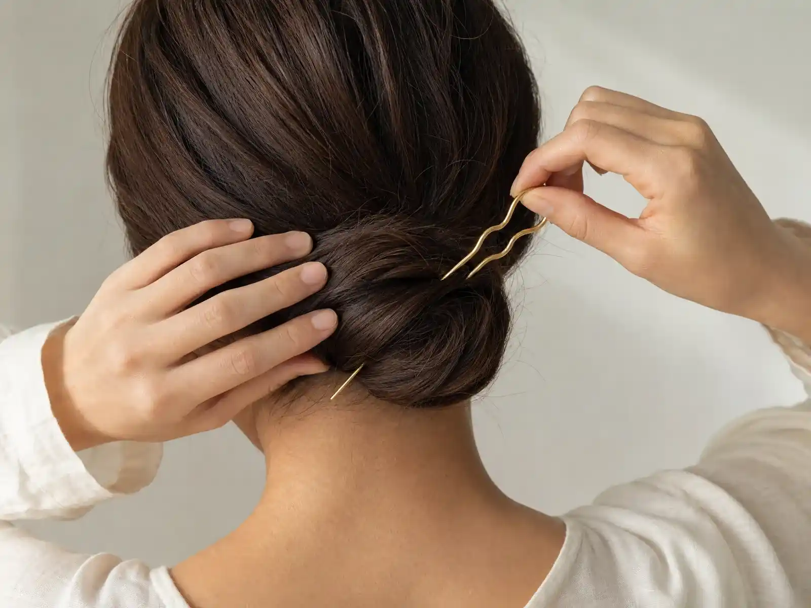Two hands setting a wavy gold French hair pin into a smooth low chignon of dark brown hair, the pin caught partway through its rotation into the base of the bun