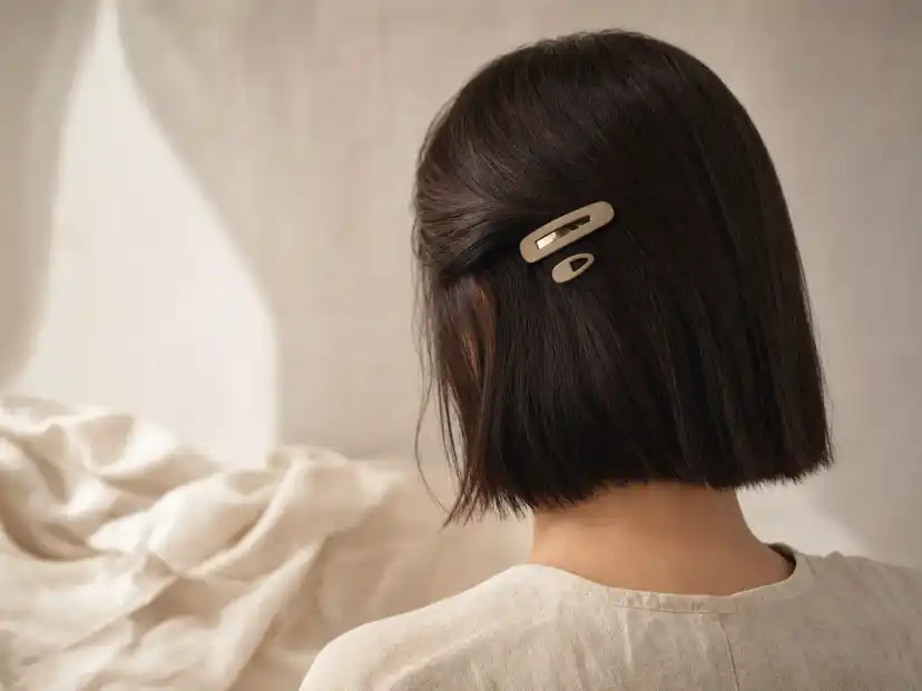 A chin-length dark brown blunt bob seen from behind and to the side, with one side swept back and held close to the head by a small matte snap barrette and a second tiny clip below it
