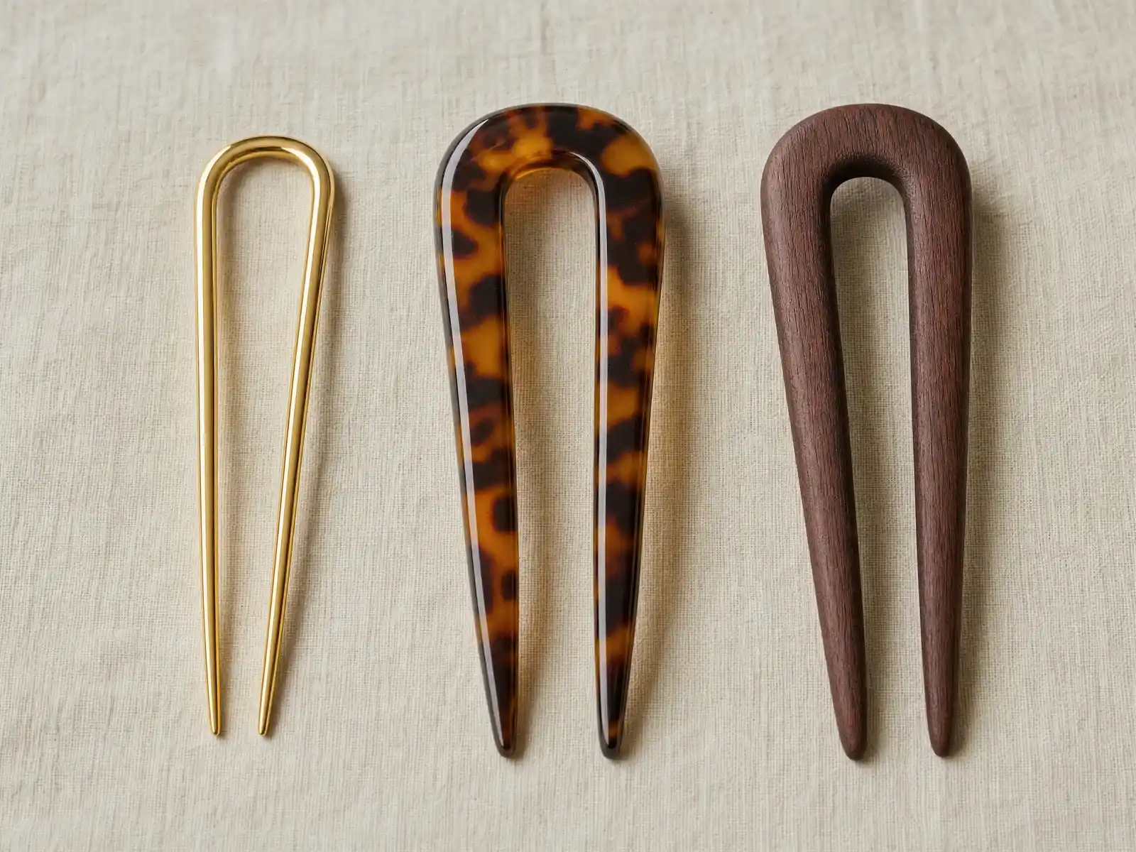 A polished gold metal French hair pin, a glossy tortoiseshell acetate pin and a matte rosewood pin laid side by side on neutral linen, showing the difference in thickness and surface finish