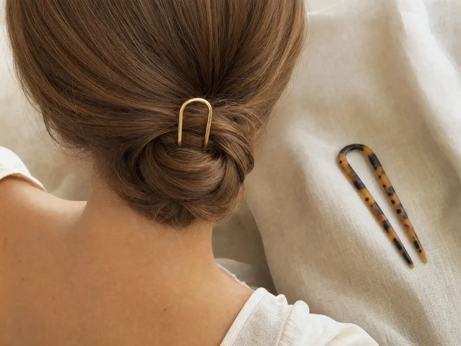 A single gold U-shaped French hair pin set into a low twisted chignon at the nape of the neck on light brown hair, with a tortoiseshell acetate hair fork resting on pale linen beside it