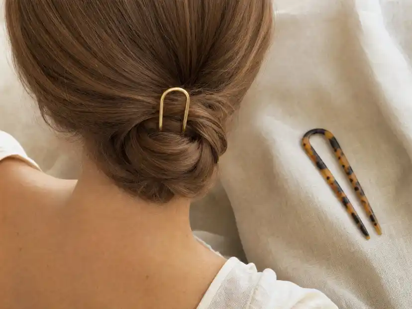 A single gold U-shaped French hair pin set into a low twisted chignon at the nape of the neck on light brown hair, with a tortoiseshell acetate hair fork resting on pale linen beside it