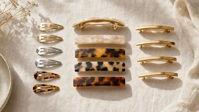 Seven types of barrettes arranged on linen, including a tortoiseshell French barrette with its tension-bar clasp open, small metal snap clips, an acetate rectangle, and a banana clip