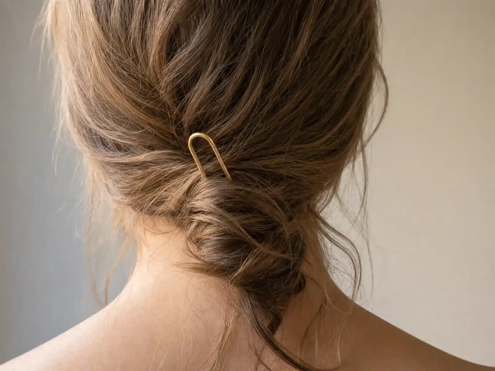 A gold U-shaped French hair pin backed halfway out of a sagging low twist of light brown hair, one prong no longer buried in the twist and loose strands falling free at the nape